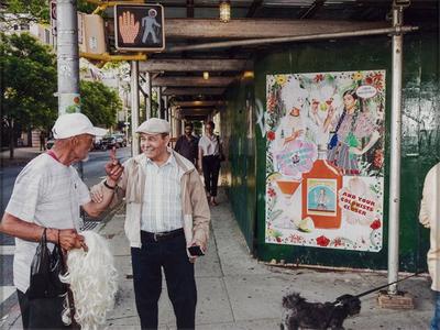busy street scene; two men in conversation in LLQ, left figure holds black shopping bag and wig with white hair in PR arm; PL hand raised in the air, gripped by right figure, who wears a tan jacket and hat, with gray mustache and smiling; covered sidewalk in center with two figures walking under covering, female figure in right wearing black pants, glasses and white striped shirt, and is talking to a male figure wearing glasses, plaid button up shirt and baseball hat; colorful advertising sign on right depicting two costumed women, saluting with drinks; traffic crossing sign in ULC