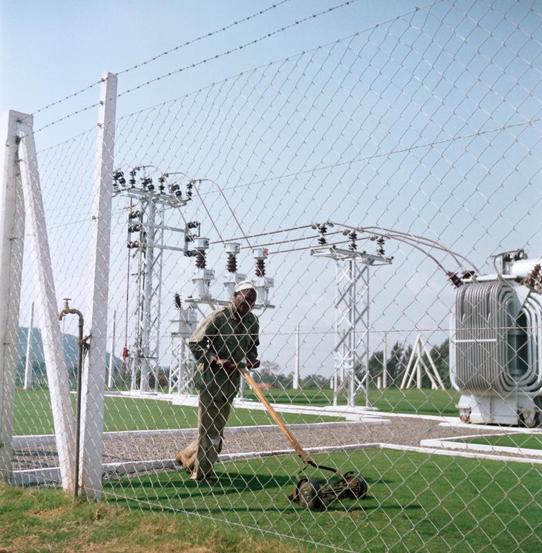 Color photograph of a man using a push-type mower to mow grass inside a fence with electrical towers behind him