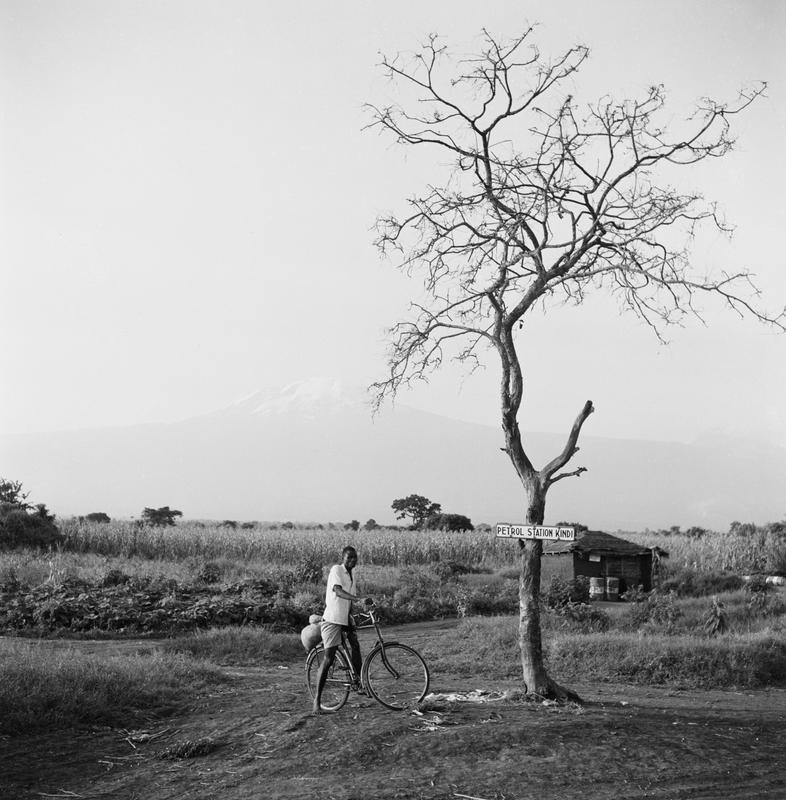 Black and white image of a man on a bike under a tree with a sign on it that says "Petrol Station Kindi"